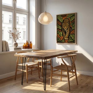 May include: A dining room scene featuring a wooden table with black hairpin legs and four wooden chairs. A framed artwork of a jaguar in a jungle setting hangs on the wall. A light fixture hangs above the table, and a window is visible.