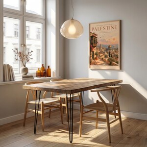 May include: A rectangular wooden dining table with black metal hairpin legs, accompanied by three light-colored wooden chairs with woven seats. A framed poster titled PALESTINE hangs on the wall, and a glass pendant light hangs above.