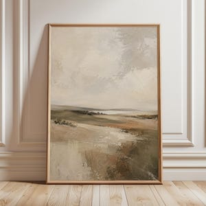 May include: A framed landscape painting in a neutral colour palette. The artwork depicts a field with muted green and brown tones, a light-coloured sky, and a distant body of water. The painting is in a wooden frame and leans against a white wall.