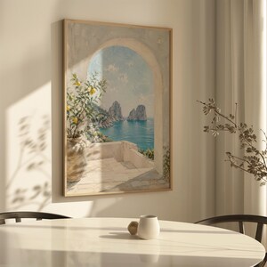 May include: A framed painting depicts a coastal scene with an archway overlooking the sea. A lemon tree in a pot is on the left. The painting has a light wooden frame and is hanging on a white wall.