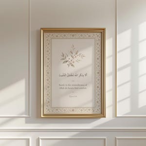 May include: A framed print with a gold-coloured border, displaying Arabic calligraphy and English text. The artwork incorporates a floral design at the top. The English text states, "Surely in the remembrance of Allah do hearts find comfort."