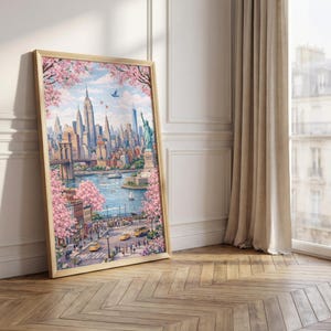 Op de afbeelding: Een ingelijste aquarelprint van New York City, met de skyline, de Brooklyn Bridge en het Vrijheidsbeeld. Roze kersenbloesems omlijsten het stadsgezicht. De print zit in een licht houten frame, leunend tegen een witte muur en een houten vloer.