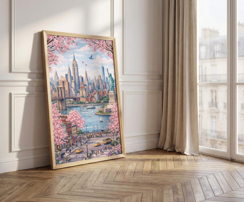 Op de afbeelding: Een ingelijste aquarelprint van New York City, met de skyline, de Brooklyn Bridge en het Vrijheidsbeeld. Roze kersenbloesems omlijsten het stadsgezicht. De print zit in een licht houten frame, leunend tegen een witte muur en een houten vloer.