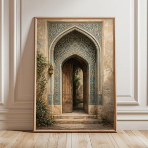 May include: A framed print depicting an arched doorway with intricate teal and gold tile patterns. The wooden door is ajar, offering a view of a garden. The artwork is in a wooden frame, resting against a white wall and wooden floor.