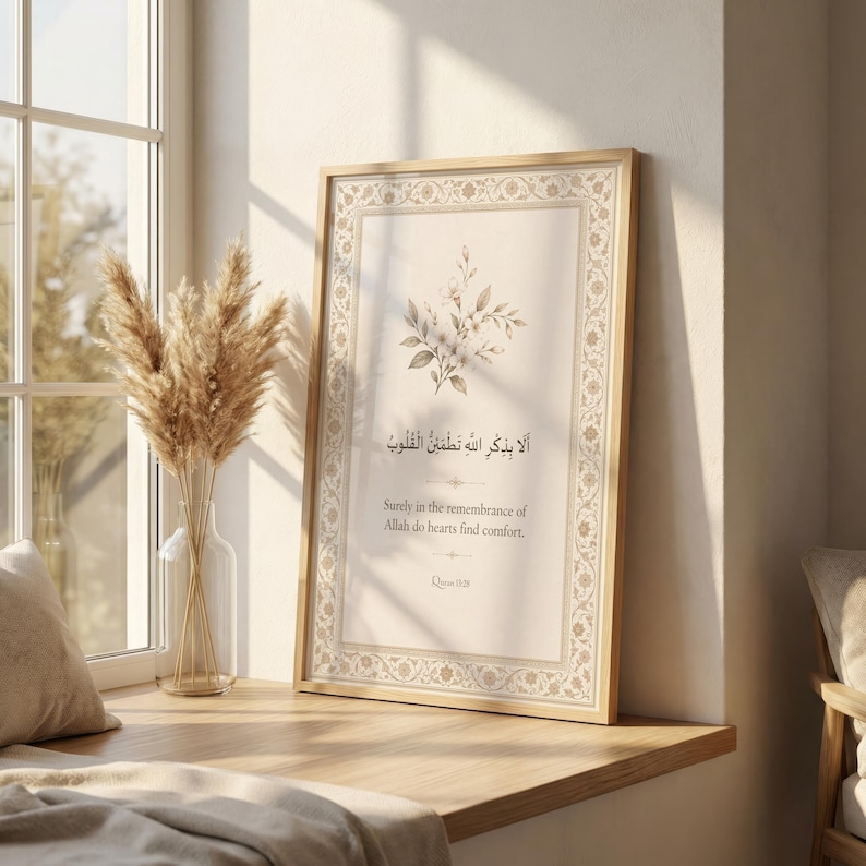 May include: A framed print with Arabic calligraphy and English translation. The artwork features floral accents and a decorative border. The frame is a light wood colour. The print is displayed on a wooden ledge near a window.