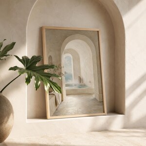 May include: A framed print showing a tranquil architectural scene with arched doorways and a coastal view. The artwork uses a muted palette of beige, white, and blue, and has a wooden frame. A potted plant with large green leaves is in the foreground.