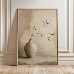May include: A framed wall art print featuring a textured, beige ceramic vase with a rounded body and a narrow neck. A single, slender branch with small, pointed leaves extends from the vase. The background is a neutral, mottled beige wall.