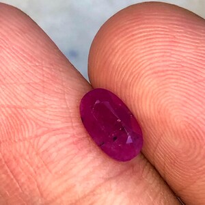 May include: A faceted, oval-cut ruby gemstone. The ruby is a deep red colour with a polished surface. The gemstone is held between two fingers, highlighting its size and clarity.