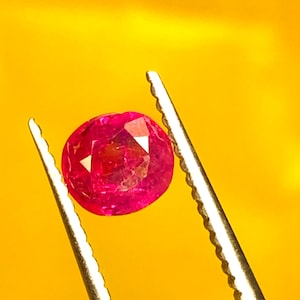 Puede incluir: Una gema de rubí redonda y facetada, sujeta por unas pinzas plateadas sobre un fondo amarillo. El rubí es de un color rojo-rosado vibrante, con facetas internas visibles que reflejan la luz. Las pinzas tienen bordes dentados.