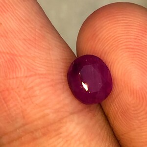 May include: A faceted, oval ruby gemstone. The gem is a deep red colour with a high degree of clarity. The ruby is held between two fingers, showcasing its polished surface and brilliance.