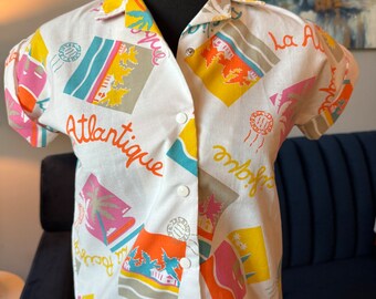 May include: A white short-sleeved button-up shirt with a colorful print of palm trees, houses, and the words "La Atlantique", "La Pacifique", and "La Alque",  "Emboffs" and "Beso My"