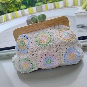 Può includere: Una pochette all'uncinetto realizzata con quadrati di filato color bianco sporco con motivi floreali pastello in giallo, rosa, blu e verde. La borsa presenta una chiusura a telaio in legno chiaro sormontata da due perline geometriche in silicone verde salvia.