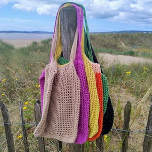 May include: A collection of colorful, open-weave crochet tote bags in various shades, including beige, purple, yellow, orange, green, and black. The bags are hanging on a wooden post with a beach and grassy dunes in the background.