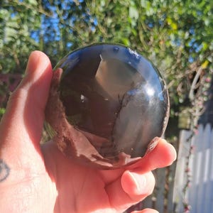 May include: A polished, dark gray crystal sphere. The sphere is held in a hand, reflecting light and the surrounding environment. The crystal ball has a smooth, rounded shape, and is a decorative item.
