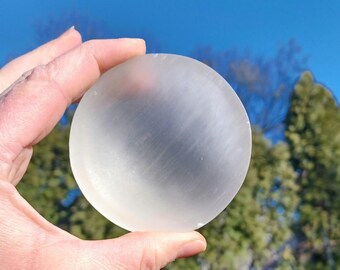 May include: A clear, white selenite sphere, also known as a moonstone, is held in a hand against a blurred background of trees and a blue sky.