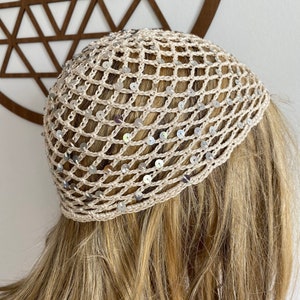 May include: A beige crocheted head covering with a net-like design, adorned with silver sequins. The head covering is worn on a person with blonde hair. The item is likely handmade and suitable for special occasions or fashion.
