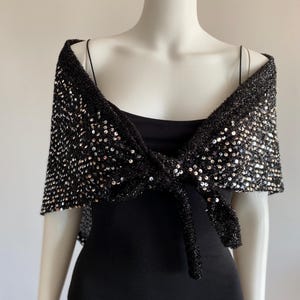 Peut inclure: Un châle en tricot noir orné d'une couverture dense de paillettes argentées réfléchissantes. Le châle présente une base en fil texturé et duveteux, noué sur le devant, se drapant sur les épaules pour offrir un aspect métallique scintillant.
