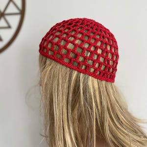 Op de afbeelding: Een felrode gehaakte hoed met een open, rasterachtig ontwerp. De hoed is gemaakt van dik garen. De hoed wordt op het hoofd van een persoon gedragen, met blond haar eronder zichtbaar.