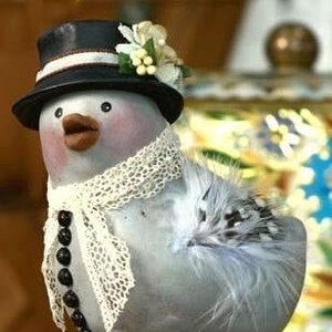 May include: A whimsical bird figurine with a black top hat, a white lace scarf, and a beaded necklace. The bird has a gray body with feather accents and stands on thin, brown legs atop a green base. Decorative floral accents adorn the hat.