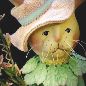 May include: A decorative bunny figurine wearing a straw hat with a pale green trim and a green leaf collar. The bunny has rosy cheeks, black eyes, and long white whiskers. It holds a bouquet of pink flowers. A gold medallion is visible.
