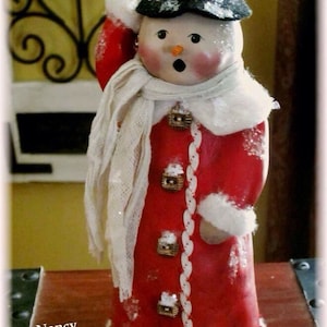 May include: A handmade snowman figurine wearing a red coat, white fur trim, a white scarf, and a black top hat with a red band. The snowman has a painted face with rosy cheeks and a surprised expression. The figurine is decorated with white glitter and has a white base.