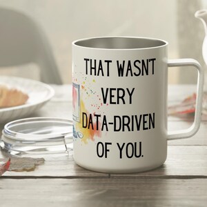 Peut inclure: Mug de voyage blanc avec un bord et une poignée argentés. Le mug porte l'inscription en noir : "THAT WASN'T VERY DATA-DRIVEN OF YOU." avec des éclaboussures d'aquarelle colorées. Un croissant et une théière sont en arrière-plan.
