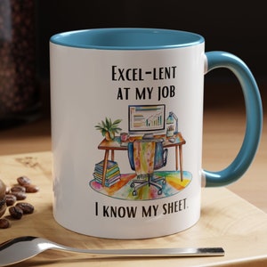 May include: White ceramic mug with a blue rim and handle. The mug features a black and white illustration of a desk with a computer, books, and a plant. The text on the mug reads "Excel-lent at my job I know my sheet."