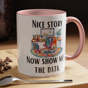 May include: White ceramic mug with a pink interior and handle. The mug features the text "Nice Story Now Show Me The Data." and a watercolour illustration of a desk with a computer, chair, and rug.