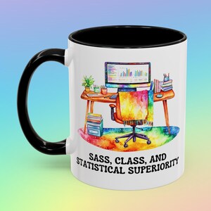 May include: White ceramic mug with a black rim and handle. The mug features a watercolor illustration of a desk with a computer, books, and a rainbow-colored chair. The text "SASS, CLASS, AND STATISTICAL SUPERIORITY" is printed on the mug.