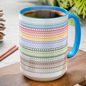 May include: A 15 ounce light blue coffee mug with a rainbow pattern of colorful lines and geometric shapes. The mug has a white interior and a light blue rim.