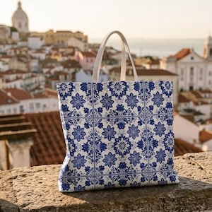 Op de afbeelding: Een tote bag met een blauw en wit bloemen tegelpatroon, met witte handvatten. De tas staat tegen een achtergrond van een stadsgezicht met rode pannendaken en een kerk. Een potplant met roze bloemen staat links.