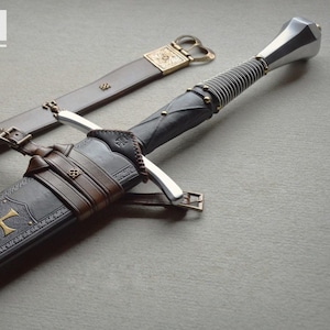 May include: A medieval-style sword with a silver blade and a black handle wrapped with wire. The sword is in a black sheath with a gold cross emblem and brown leather straps. The text "BLAYDZ_KNIFE_CO" is at the top.