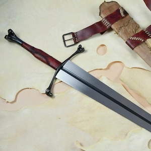 May include: A long, silver sword with a dark red handle and crossguard, alongside a leather sheath and belt. The sheath is tan and brown, with stitched accents. The text "BLAYDZ_KNIFE_CO" is visible.