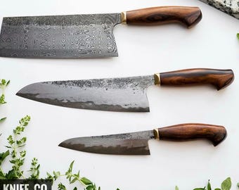 May include: Three handcrafted kitchen knives with dark wooden handles and textured blades are displayed on a white surface. The knives vary in size, with a cleaver-style knife at the top. The brand name "BLAYDZ_KNIFE.CO" is visible at the bottom.