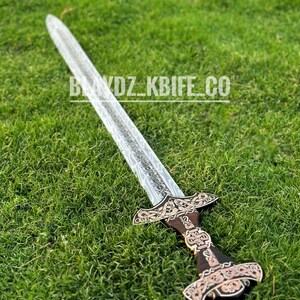May include: A silver sword with a wooden handle and intricate carvings. The sword is lying on a bed of green grass.