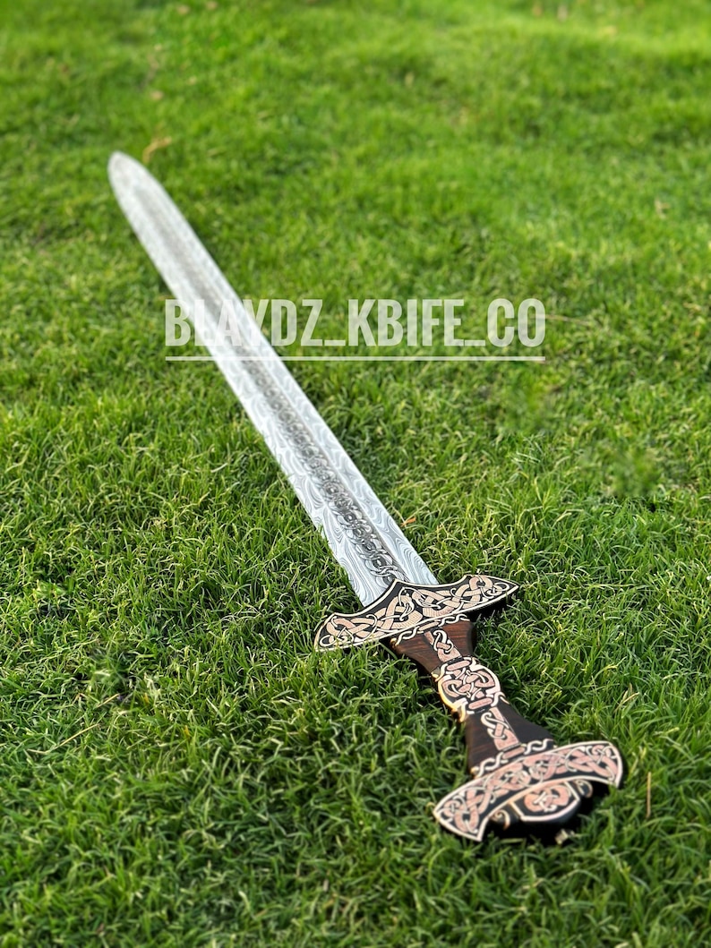 May include: A silver sword with a wooden handle and intricate carvings. The sword is lying on a bed of green grass.