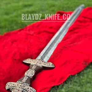 May include: A silver sword with a dark wood handle and intricate metalwork on the pommel and guard. The sword is lying on a red cloth and a green grassy background. The text "BLAYDZ_KNIFE_CO" is visible in the image.