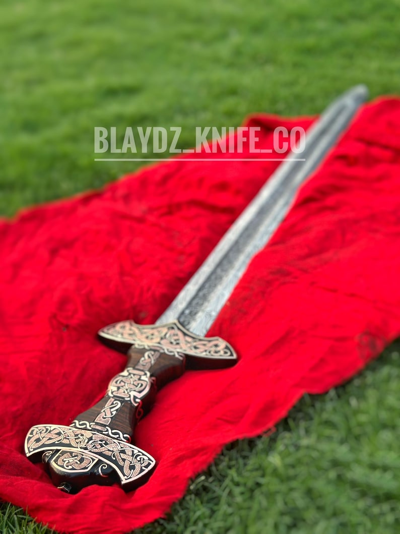 May include: A silver sword with a dark wood handle and intricate metalwork on the pommel and guard. The sword is lying on a red cloth and a green grassy background. The text "BLAYDZ_KNIFE_CO" is visible in the image.
