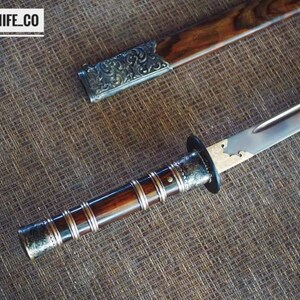 May include: A close-up of a sword with a wooden handle and a silver blade. The handle is dark brown with silver detailing. The scabbard is also visible, with a wooden body and silver detailing. The text "BLAYDZ_KNIFE_CO" is in the top left corner.