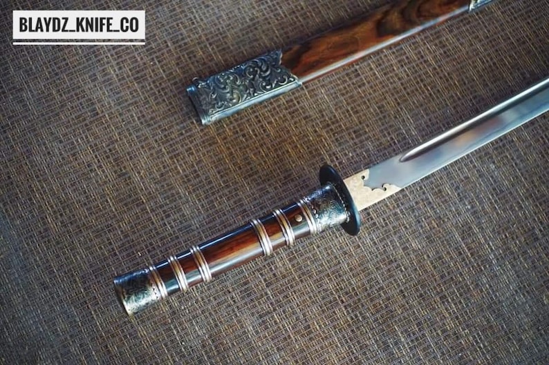 May include: A close-up of a sword with a wooden handle and a silver blade. The handle is dark brown with silver detailing. The scabbard is also visible, with a wooden body and silver detailing. The text "BLAYDZ_KNIFE_CO" is in the top left corner.