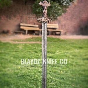 May include: A silver sword with a copper-colored hilt and intricate carvings. The sword has a long, straight blade and a cross-shaped guard. The sword is standing upright on a green grassy surface. The text "BLAYDZ_KNIFE_CO" is visible in the lower center of the image.