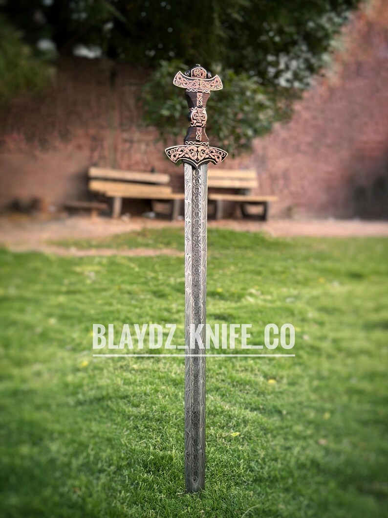 May include: A silver sword with a copper-colored hilt and intricate carvings. The sword has a long, straight blade and a cross-shaped guard. The sword is standing upright on a green grassy surface. The text "BLAYDZ_KNIFE_CO" is visible in the lower center of the image.