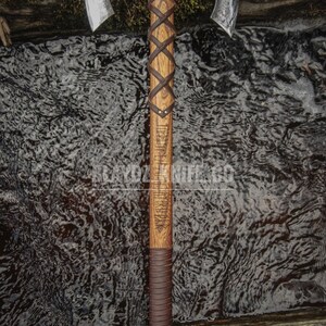 May include: A double-headed axe with a wooden handle wrapped in brown leather. The handle is carved with runes and has a leather grip. The axe is resting on a wooden surface with a stream of water in the background.