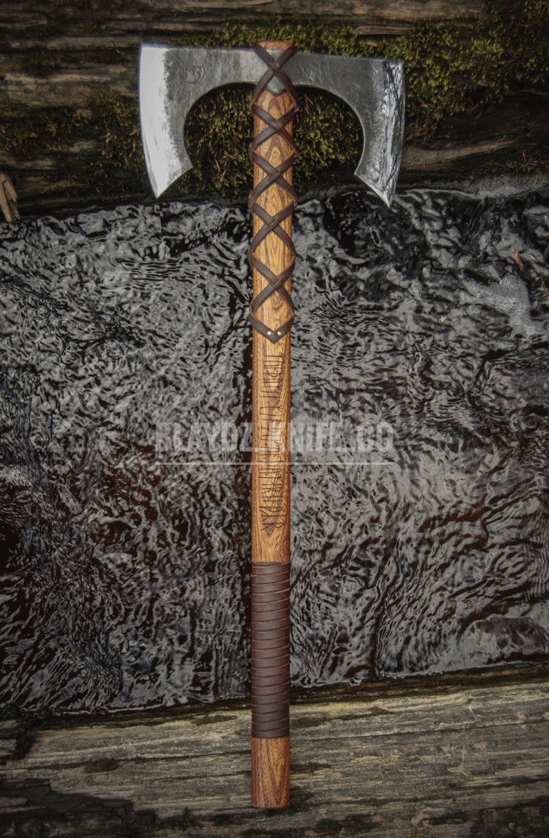 May include: A double-headed axe with a wooden handle wrapped in brown leather. The handle is carved with runes and has a leather grip. The axe is resting on a wooden surface with a stream of water in the background.