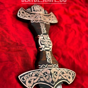 May include: A decorative sword with a black wooden handle and a silver blade. The handle is adorned with intricate copper-colored Celtic knotwork designs. The blade is Damascus steel with a wavy pattern. The sword is on a red fabric background. The text "BLAYDZ_KNIFE_CO" is visible at the top of the image.