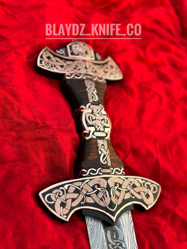 May include: A decorative sword with a black wooden handle and a silver blade. The handle is adorned with intricate copper-colored Celtic knotwork designs. The blade is Damascus steel with a wavy pattern. The sword is on a red fabric background. The text "BLAYDZ_KNIFE_CO" is visible at the top of the image.