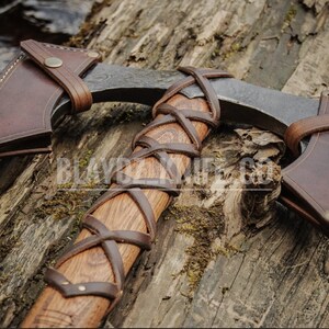 May include: A hand-forged axe with a brown leather handle wrap and sheath. The axe head is made of steel and has a curved blade. The sheath has a brass button closure.