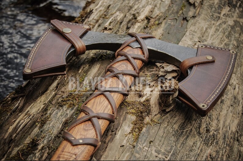 May include: A hand-forged axe with a brown leather handle wrap and sheath. The axe head is made of steel and has a curved blade. The sheath has a brass button closure.