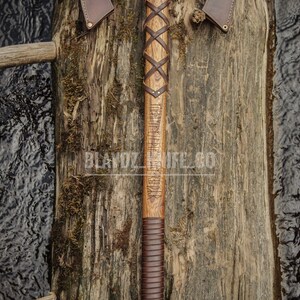 May include: A double-headed axe with a brown leather handle and a wooden shaft. The shaft is engraved with runes and the axe head has a brown leather sheath on each side. The axe is resting on a wooden surface.