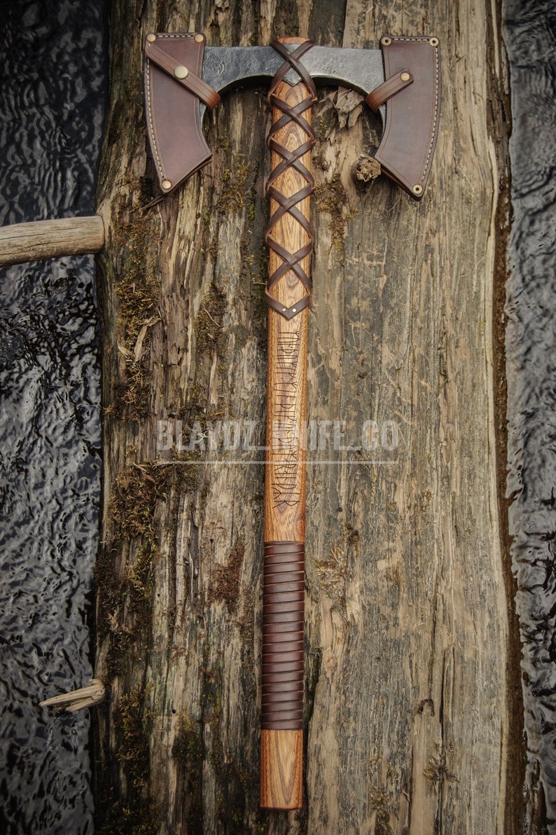 May include: A double-headed axe with a brown leather handle and a wooden shaft. The shaft is engraved with runes and the axe head has a brown leather sheath on each side. The axe is resting on a wooden surface.